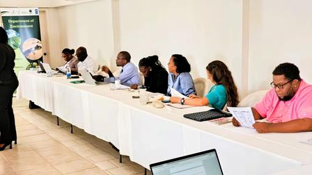 People seated along a long table with laptops in a bright conference room; presenter at left.