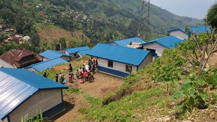 Hilly rural village with blue metal-roofed huts along a dirt road; people gather nearby.
