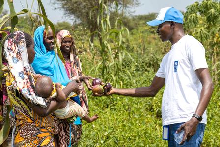 Man in white shirt and blue cap hands an item to a group of women with a baby in a rural field.