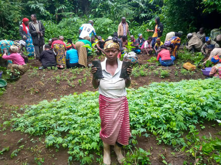 Woman in white top and pink skirt stands in a leafy garden; group gathers in a rural setting.