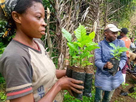 Person in gray shirt holds a potted plant as a photographer looks on in a tropical garden.