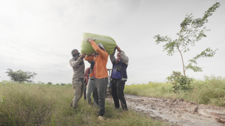 Photo shows four people carrying a large green rectangular object across a grassy rural field.