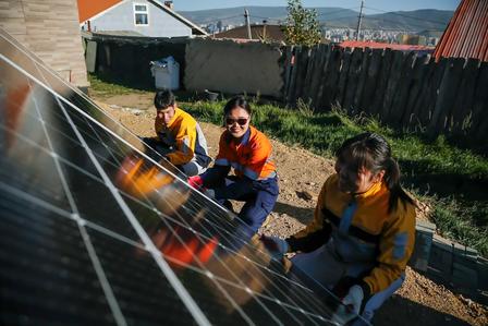 Photograph shows three workers in orange jackets kneeling beside a solar panel array.