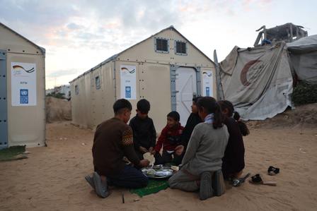 Group of people sits in a circle on sandy ground at a camp, with portable toilets in background.