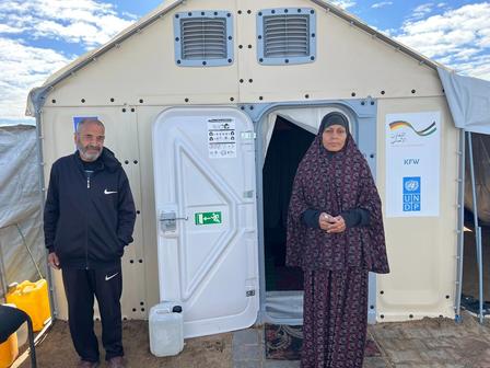 Two adults stand beside a beige portable shelter with open double doors on a sunny day.
