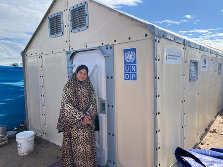 Person in a patterned robe standing at the doorway of a beige UNHCR tent against a blue sky.