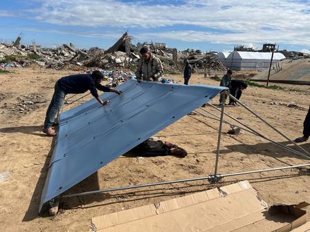 Two workers lift a large gray metal panel on a sandy construction site under a blue sky.