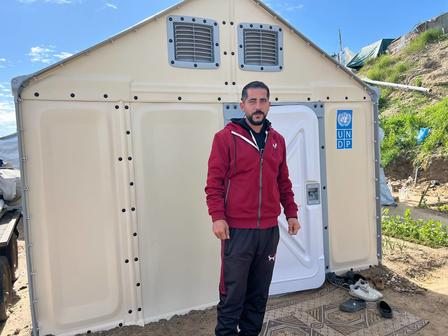 Photo: Man in red jacket standing beside a beige portable shelter outdoors.