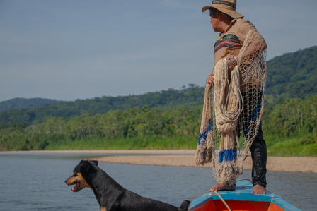 Man in a hat on a red boat by a sandy beach; a black dog swims nearby.