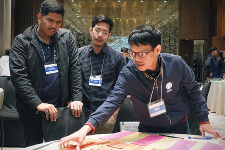Three men with badges at a conference study a colorful game board on a table, one pointing.