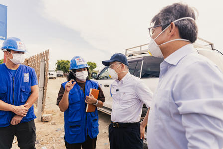 Supervisor wearing a mask talks to workers in blue uniforms and hard hats at a construction site.
