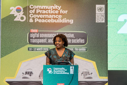 Photo: Female speaker at teal podium on stage with governance and peacebuilding conference banner.