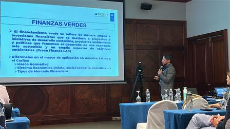 Presenter in a conference room presenting a slide titled Finanzas Verdes to an audience.