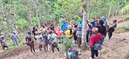 Large group of people gathered on a dirt trail in a forest clearing, many wearing backpacks.
