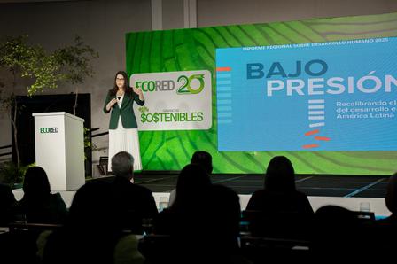 Speaker at podium on stage with a 'BAJO PRESIÓN' screen; audience silhouettes.