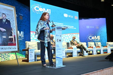 Female speaker at podium on a blue conference stage with banners and seated panelists.