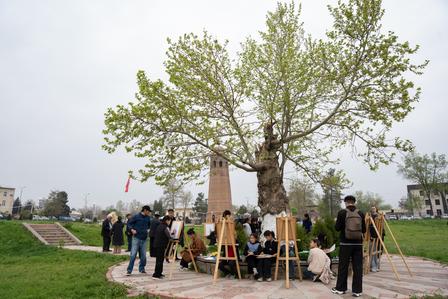 People gathered around a tall tree with a monument in a circular brick plaza at a park.