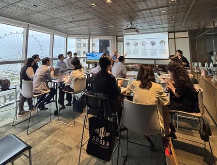 Photo: Group of adults seated around tables in a bright classroom, watching a slide presentation.