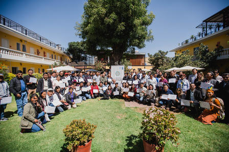 Group of people seated on a grassy courtyard circle around a tree, yellow buildings behind.