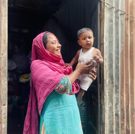 Photograph: woman in pink hijab and turquoise dress holding a baby at a wooden doorway.