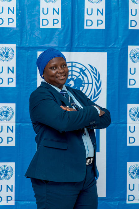 Photograph of a woman in a dark blue blazer with a lanyard, arms crossed, UNDP backdrop.
