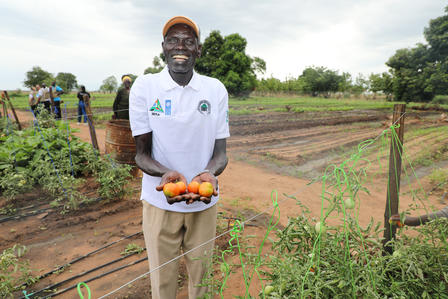 Photograph of a smiling man in a white polo and orange cap holding oranges in a farm field.