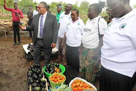 Officials in white shirts with a suited man at an outdoor market; baskets of oranges on display.