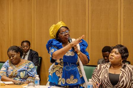 Photograph of a woman in a blue dress and yellow headwrap speaking into a microphone at a panel.
