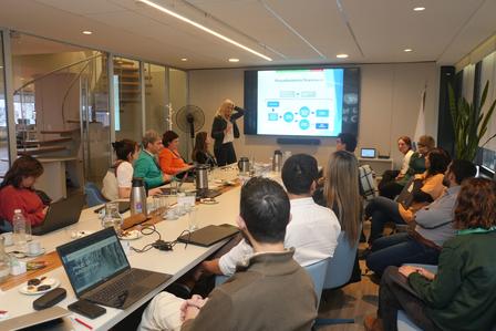Diverse group at a long conference table watches a presenter with a slide deck in a modern conference room.
