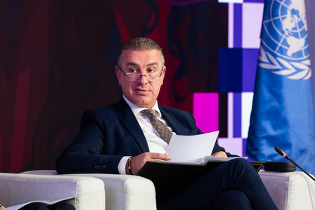 Man in suit seated on stage, reading papers; UN flag in background, face blurred.