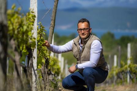 Person kneeling in a vineyard, wearing a white shirt and vest, with sea and hills behind.