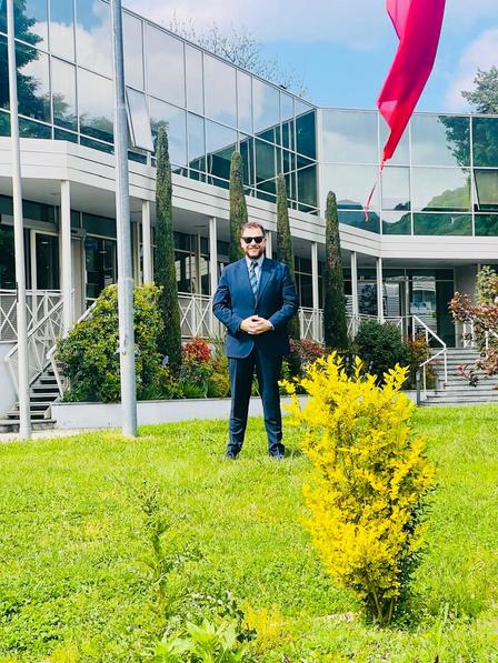 Man in a blue suit and sunglasses stands on a lawn outside glass-fronted building with red flag.