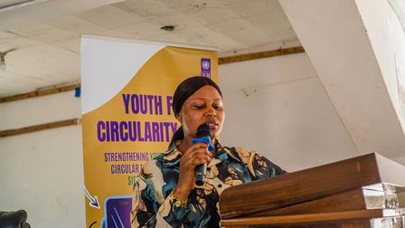 Person in a colorful patterned shirt speaks at a wooden podium; yellow and purple banner behind.
