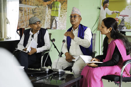 Two men in Indian attire and a woman in pink discuss around a table with microphones.
