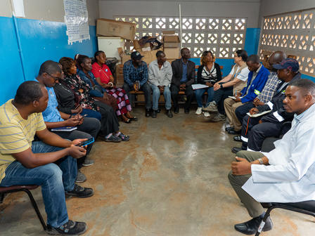 Photograph of a community meeting: people seated in a circle in a blue-walled room.