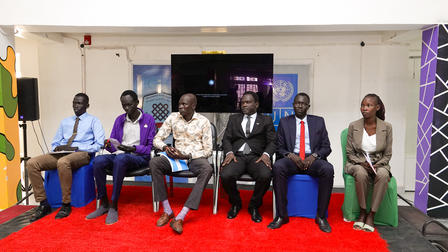 Seven people in formal attire seated on a red-carpet stage with a Nigerian flag on the left.