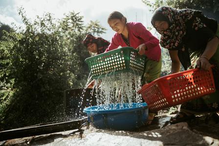 Group of people washing clothes outdoors using colorful plastic baskets by a flowing stream.