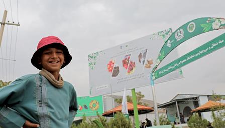 Person with blurred face wearing red cap and teal jacket at outdoor market with green banners.