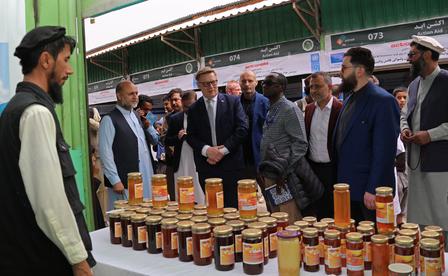 Group of men in suits around a table of honey jars at an outdoor market under a green canopy.