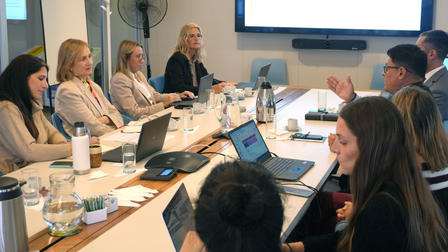 Colleagues in a conference room around a long table with laptops and a screen showing a presentation