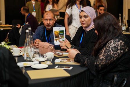 Group around a round conference table; a woman in black lace shows a phone.