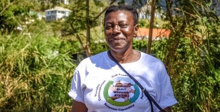 Person outdoors in white t-shirt with circular logo; blurred face, crossbody bag, green background.