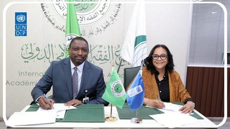Two professionals signing documents at a formal signing ceremony; banners and flags behind.