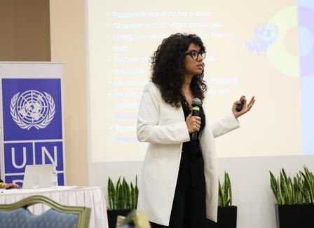 Female presenter in a white blazer speaks into a microphone on stage beside a UN banner.