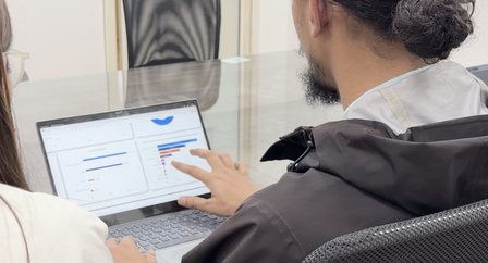 Two people sit at a desk, looking at a tablet; one person points at the screen.