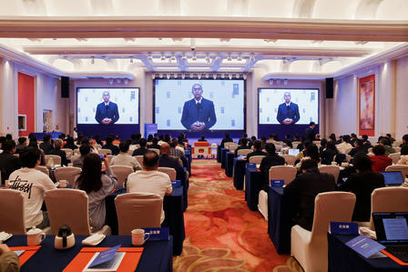 Photo: conference hall full of attendees watching a speaker on three large screens.
