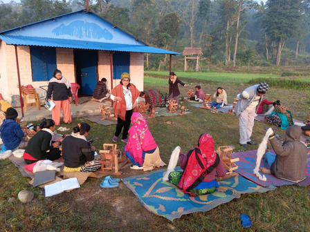 Group of people sitting on blue mats outside a small blue-roofed building in a rural setting.