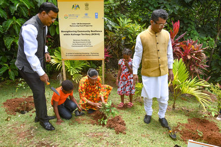Two adults and two children plant saplings in a garden beside a yellow sign.