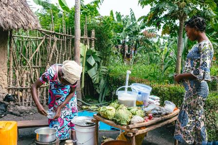 Two women in a rural yard near a thatched hut, sorting fruit at a wooden stall.