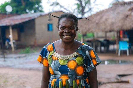 Person in a vivid patterned dress stands outdoors in a rural village with thatched huts.
